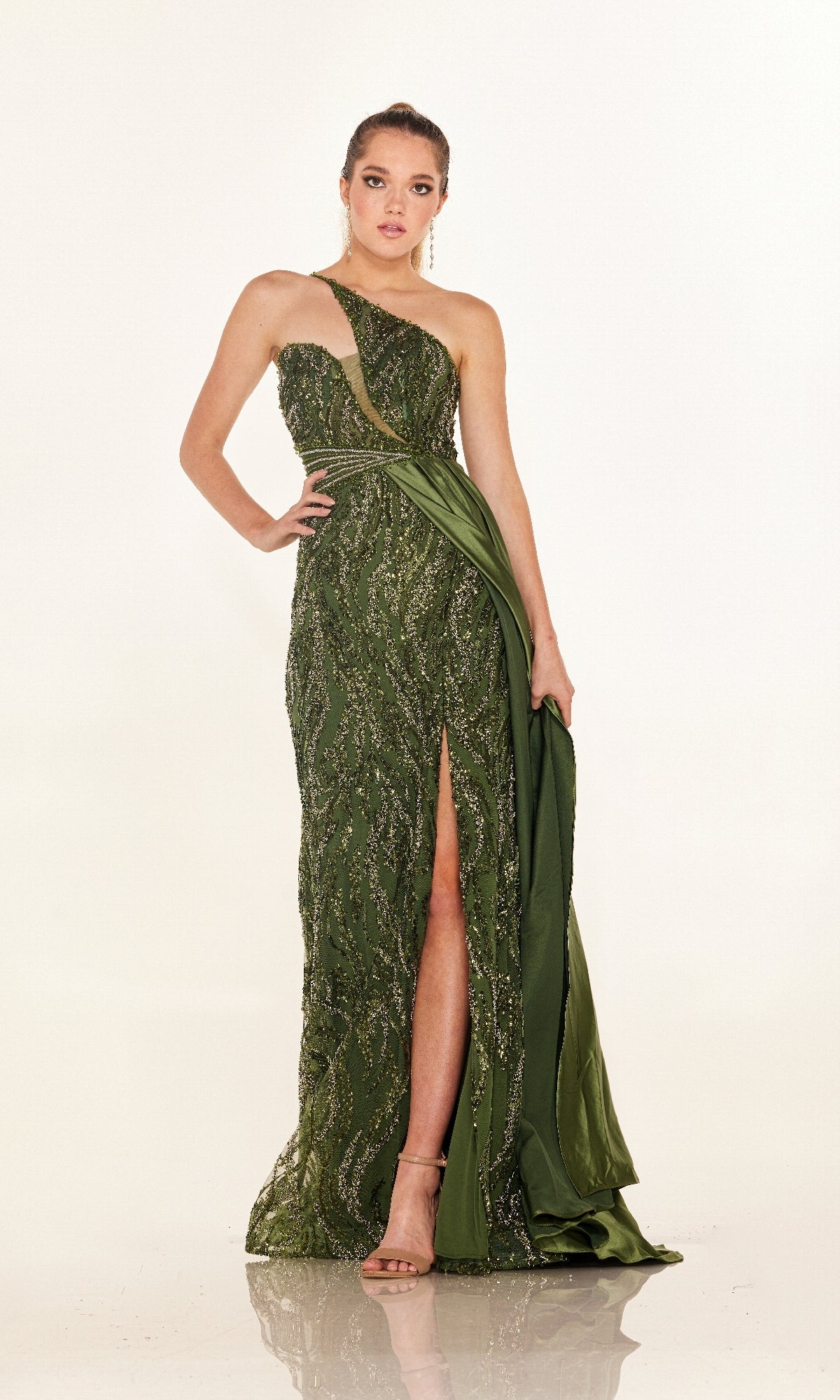 A woman stands in a studio wearing the Cecilia Couture C275, a green one-shoulder sequin long prom dress with a thigh-high slit and draped fabric, paired with beige high-heeled sandals.